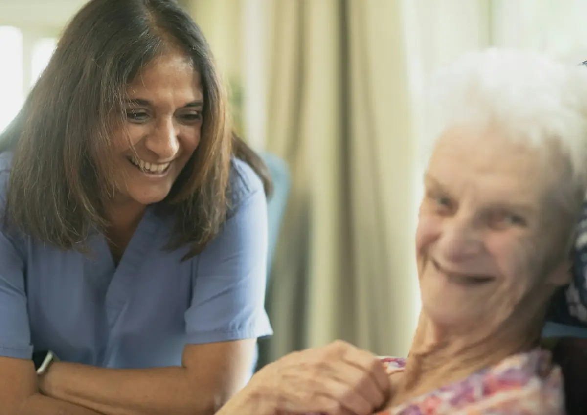 Feste Betreuerin lacht mit einer Seniorin – Hand aufs Herz Herford